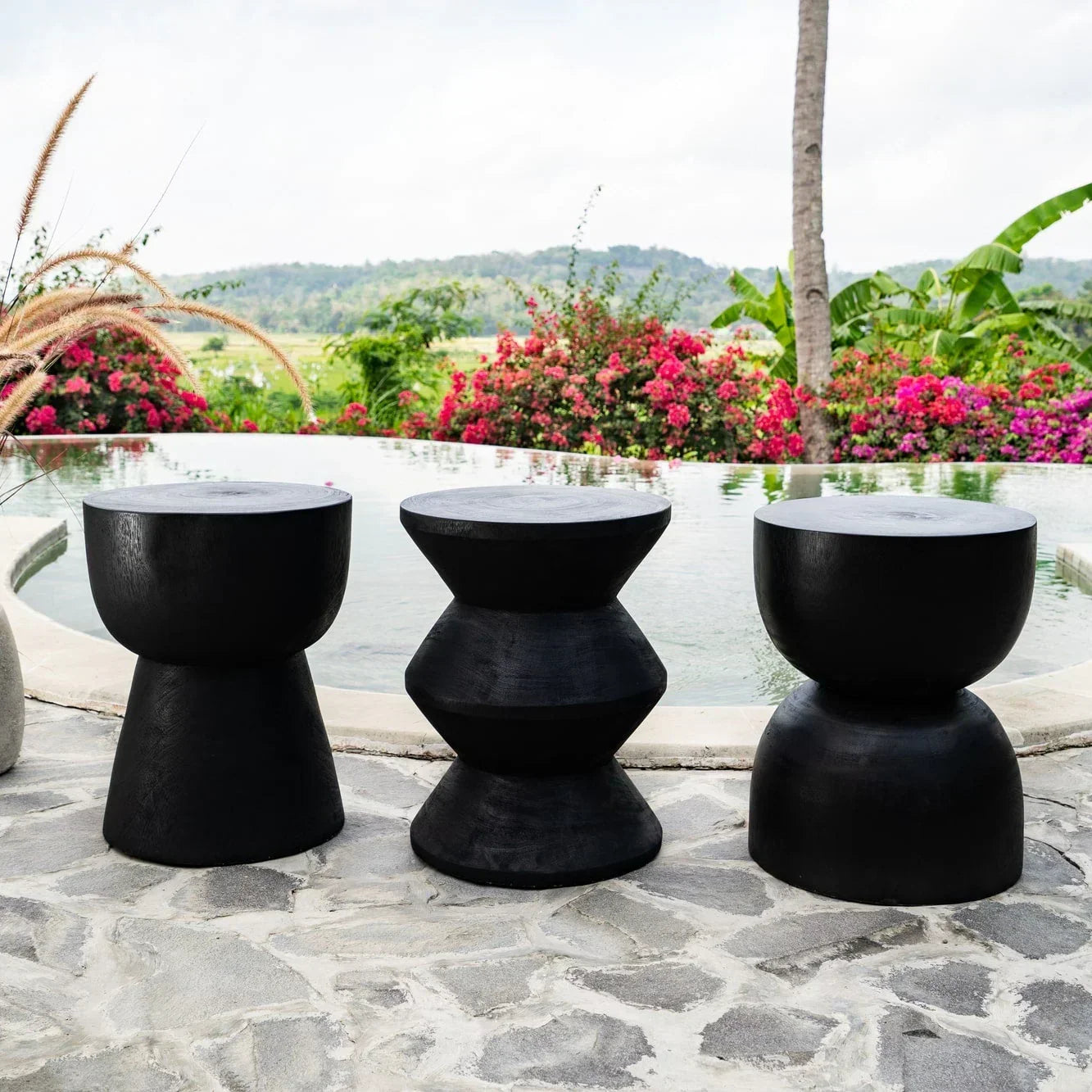 Three black hand-carved stools by a poolside with flowers and natural stone patio, Kayu Living