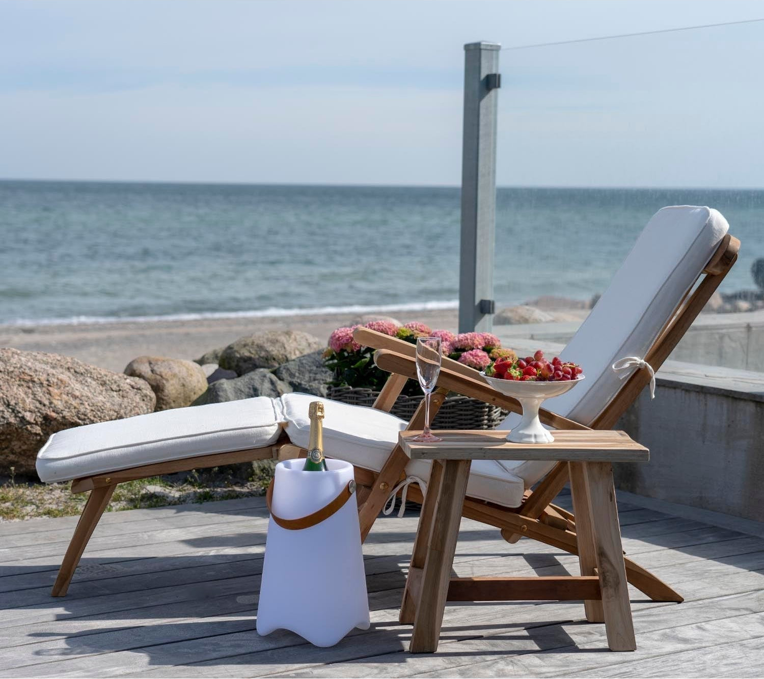 Kayu Living Arrecife teak deck chair with adjustable back and white cushion, set on a wooden deck by the sea.