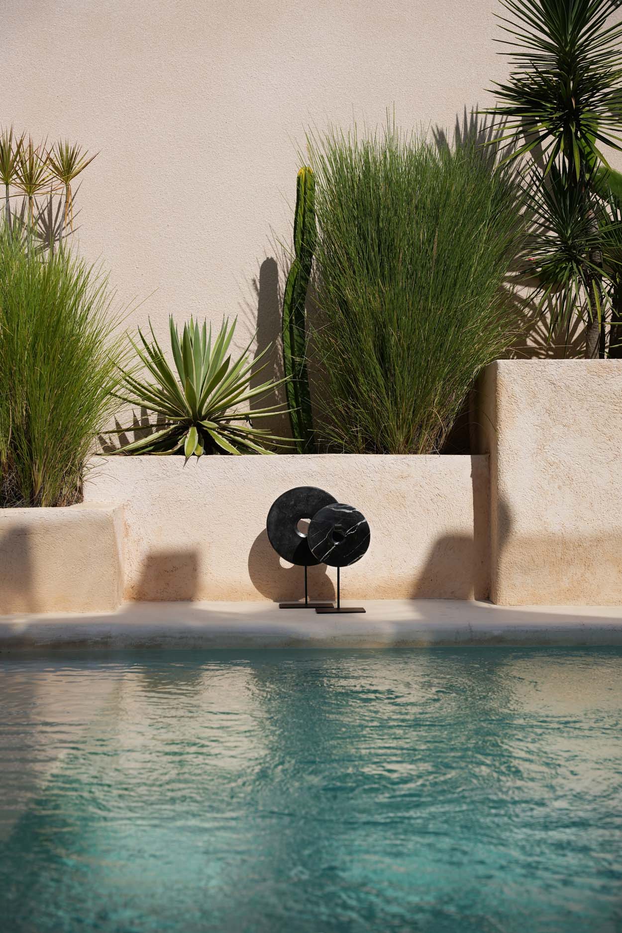 Kayu Living black marble disc sculptures on metal stands displayed by a swimming pool with desert landscaping.