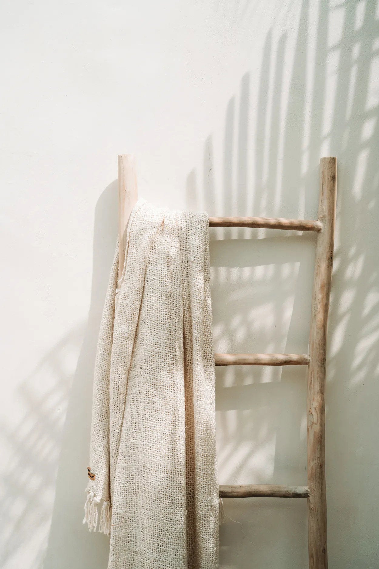 Kayu Living cream hand-woven cotton throw blanket with fringe draped over a rustic wooden ladder in sunlight