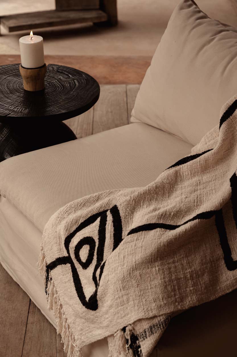 Kayu Living natural black cotton bed throw with evil eye pattern draped over a beige sofa with a candle nearby.