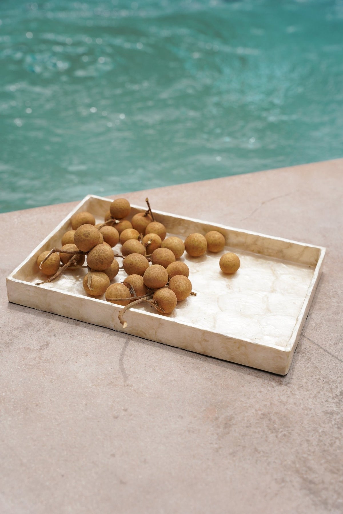 Kayu Living rectangular gold Capiz shell tray holding a cluster of longan fruit by a swimming pool