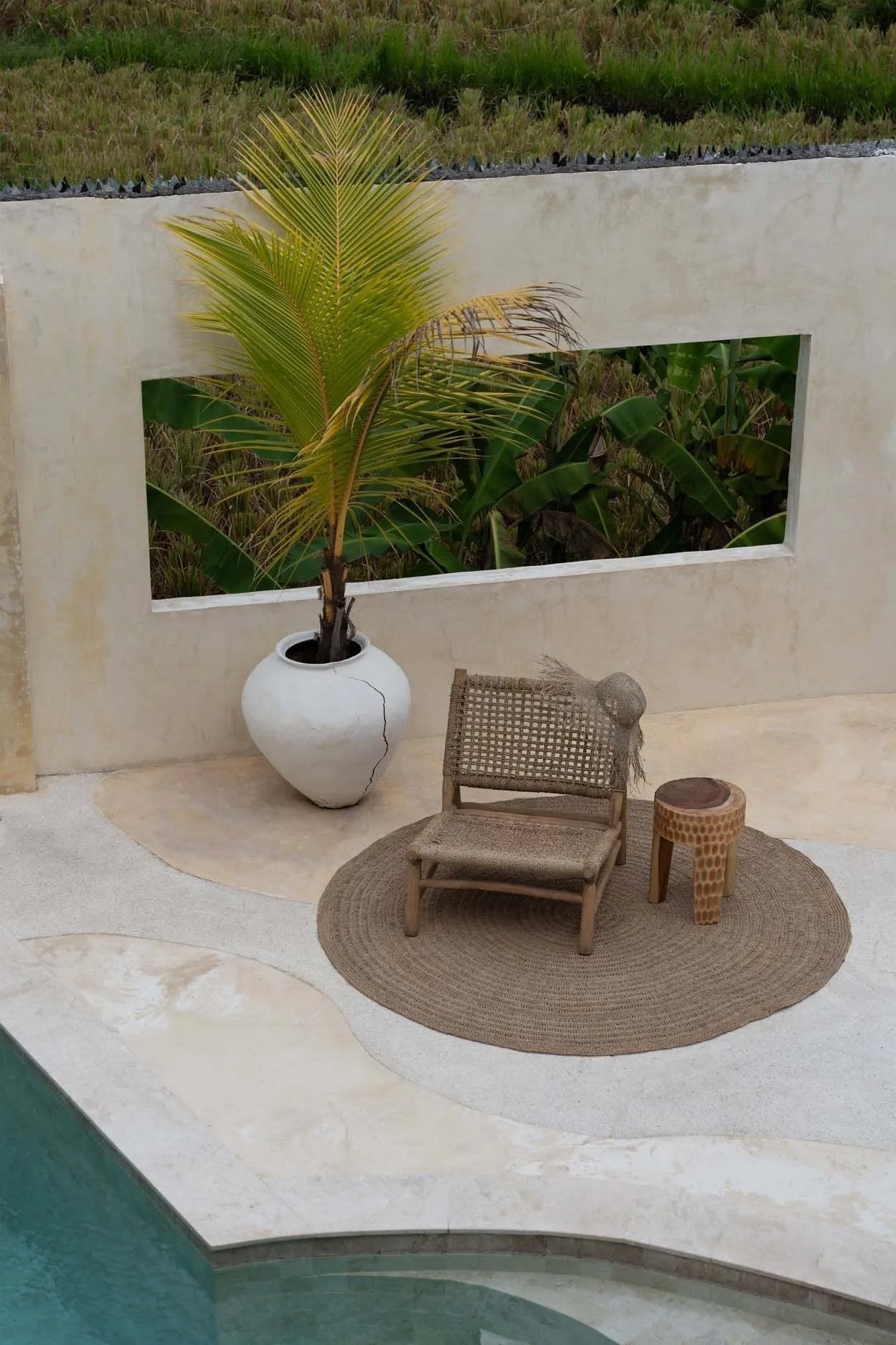 Kayu Living Paramo Stool made of natural suar wood with a three-legged minimalist design in a tropical patio setting.