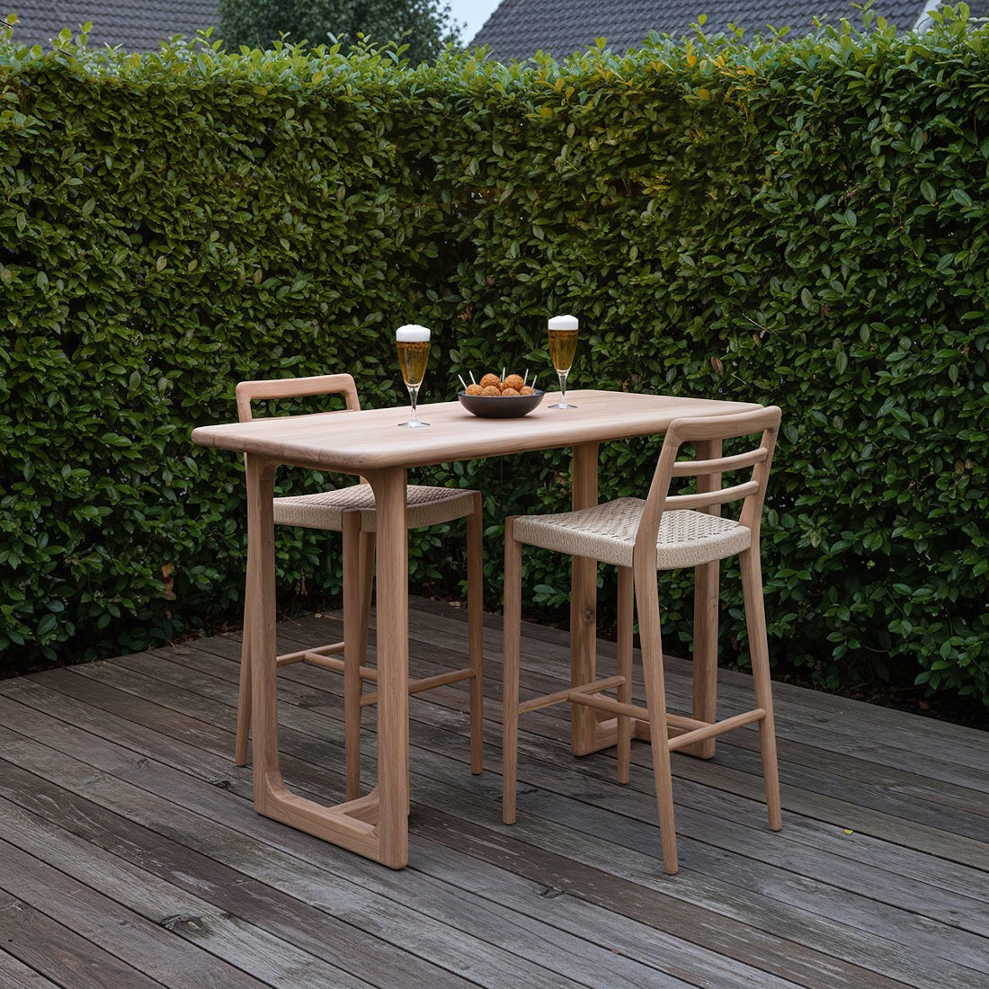 Kayu Living Primavera teak bar table with U-shaped legs and two matching stools on a wooden terrace.