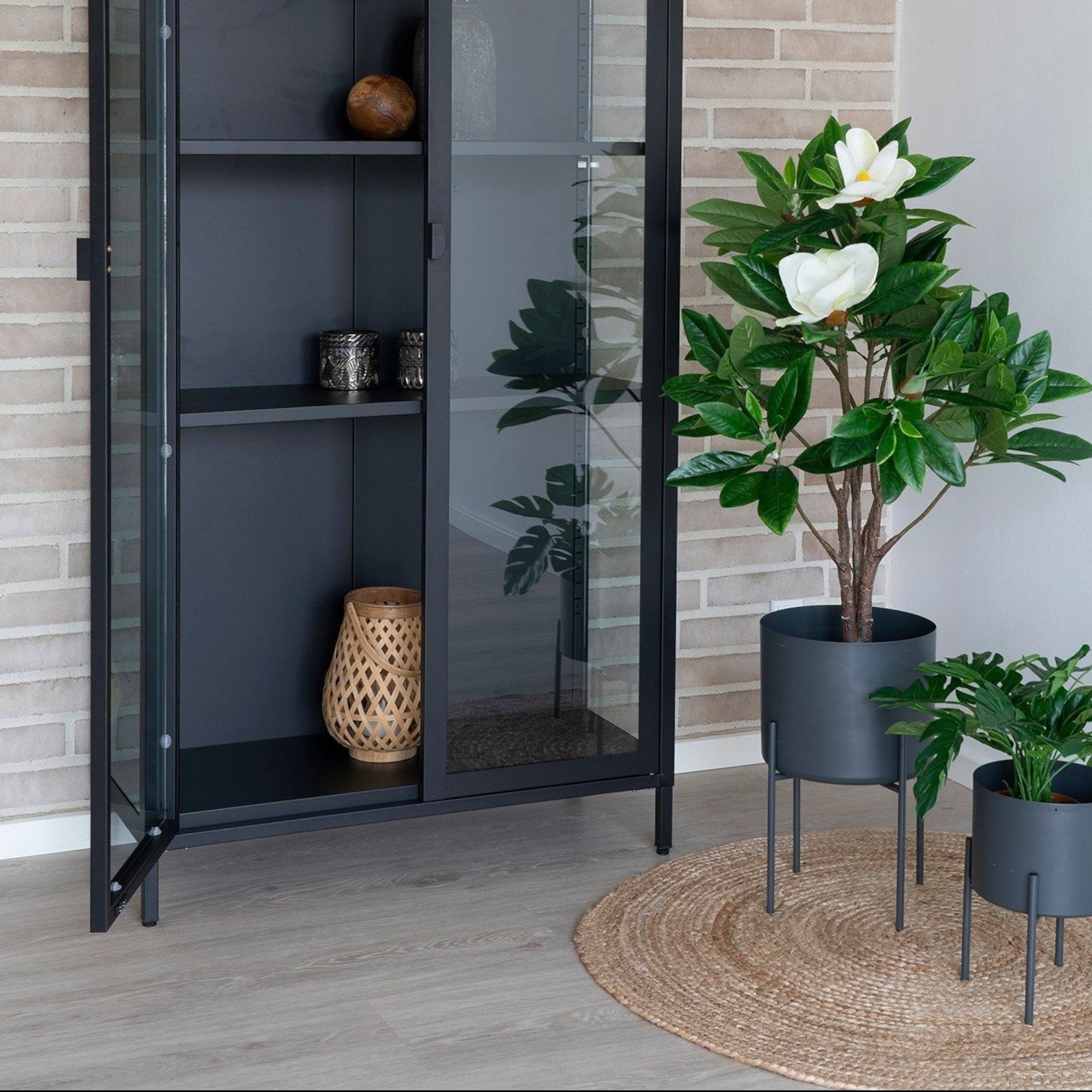Kayu Living round braided natural jute rug placed on a light wood floor next to a black display cabinet and plants.