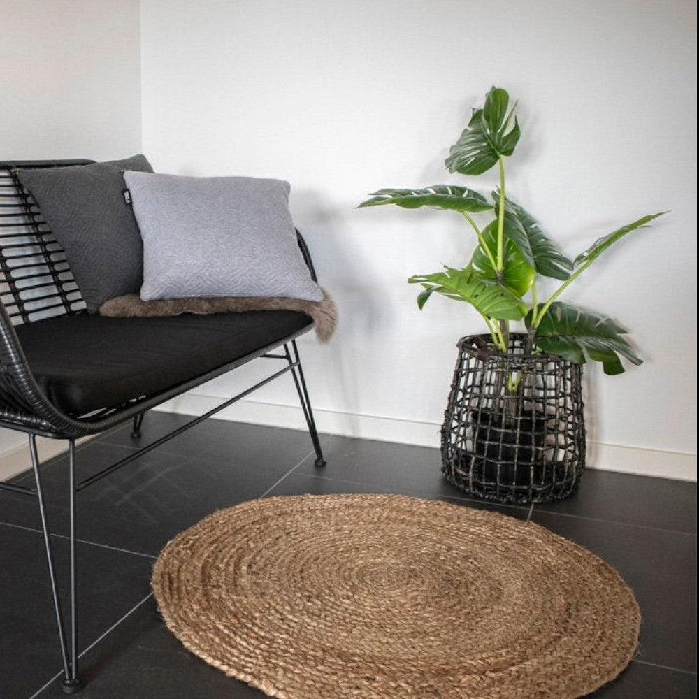 Kayu Living round natural braided jute rug placed on a dark floor next to a black rattan bench and a potted plant.
