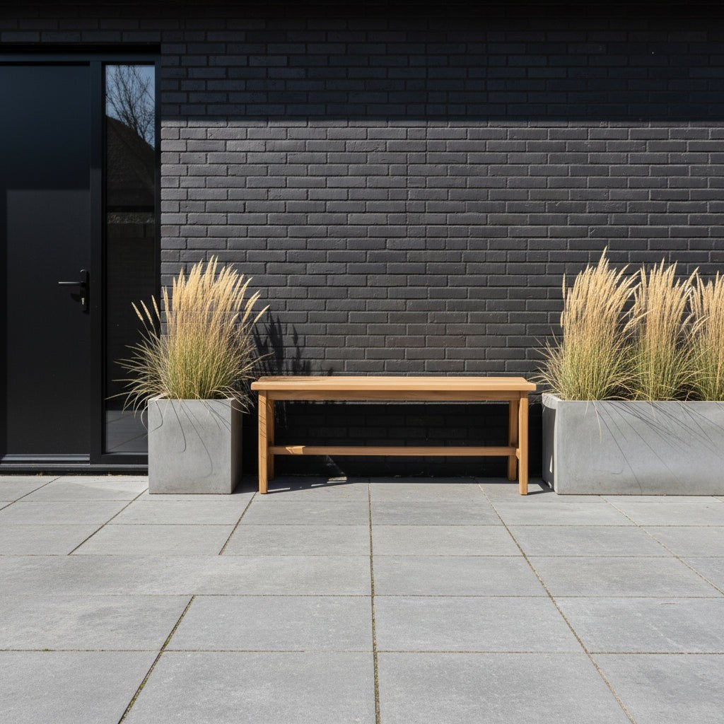 Kayu Living sustainable teak wood bench on a grey stone patio against a modern black brick wall with tall grasses.