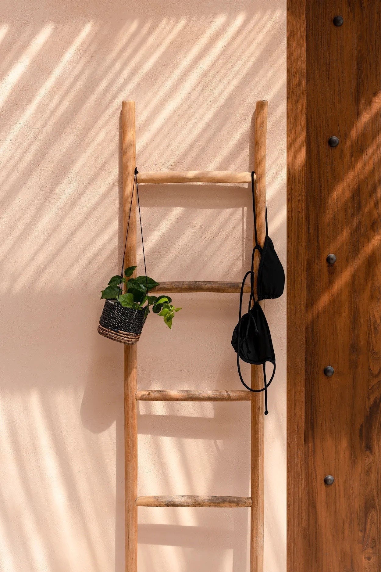 Kayu Living Tulum teak ladder leaning against a beige wall with a hanging plant and black fabric accessory