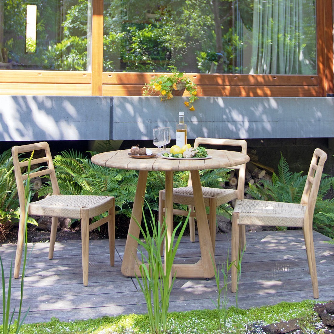 Kayu Living Verano round teak table with woven chairs on a wooden patio, perfect for outdoor dining.
