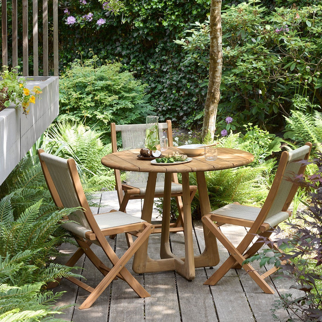 Kayu Living Verano round teak dining table with three folding chairs on a wooden patio in a lush garden setting