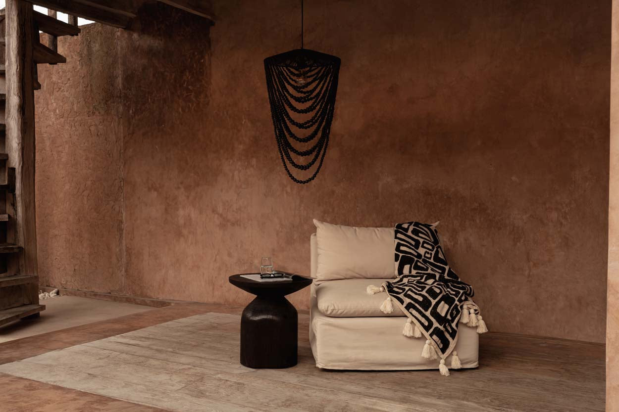 Kayu Living Aztec-pattern cotton throw in natural black with tassels draped over a beige armchair in a rustic room.