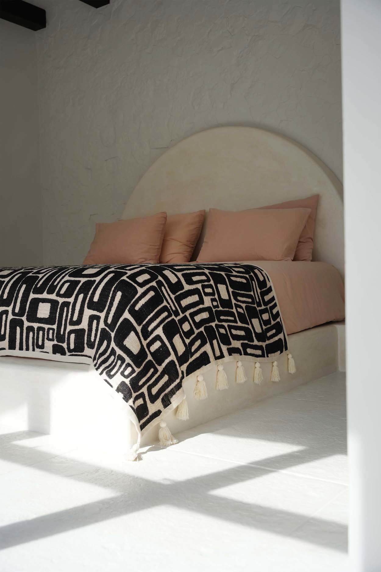 Kayu Living Aztec-pattern cotton bed throw in natural black with tassels draped over a bed with blush pink pillows