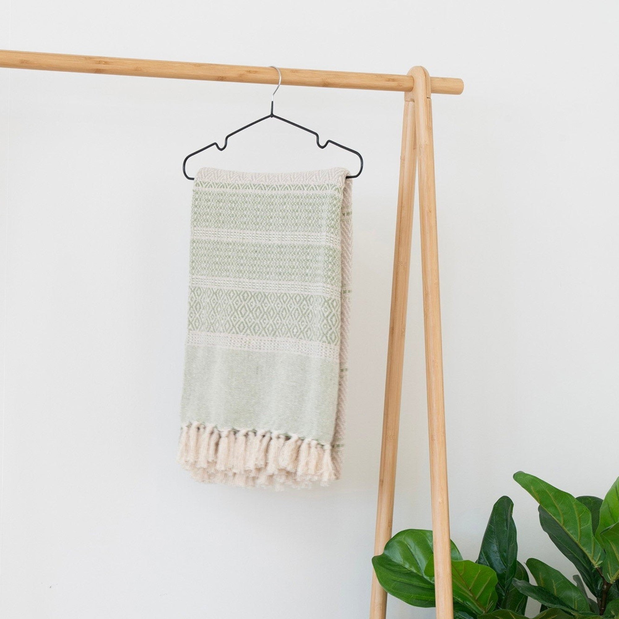 Kayu Living natural bamboo clothes rack with a green and cream patterned throw blanket with fringe hanging on it.