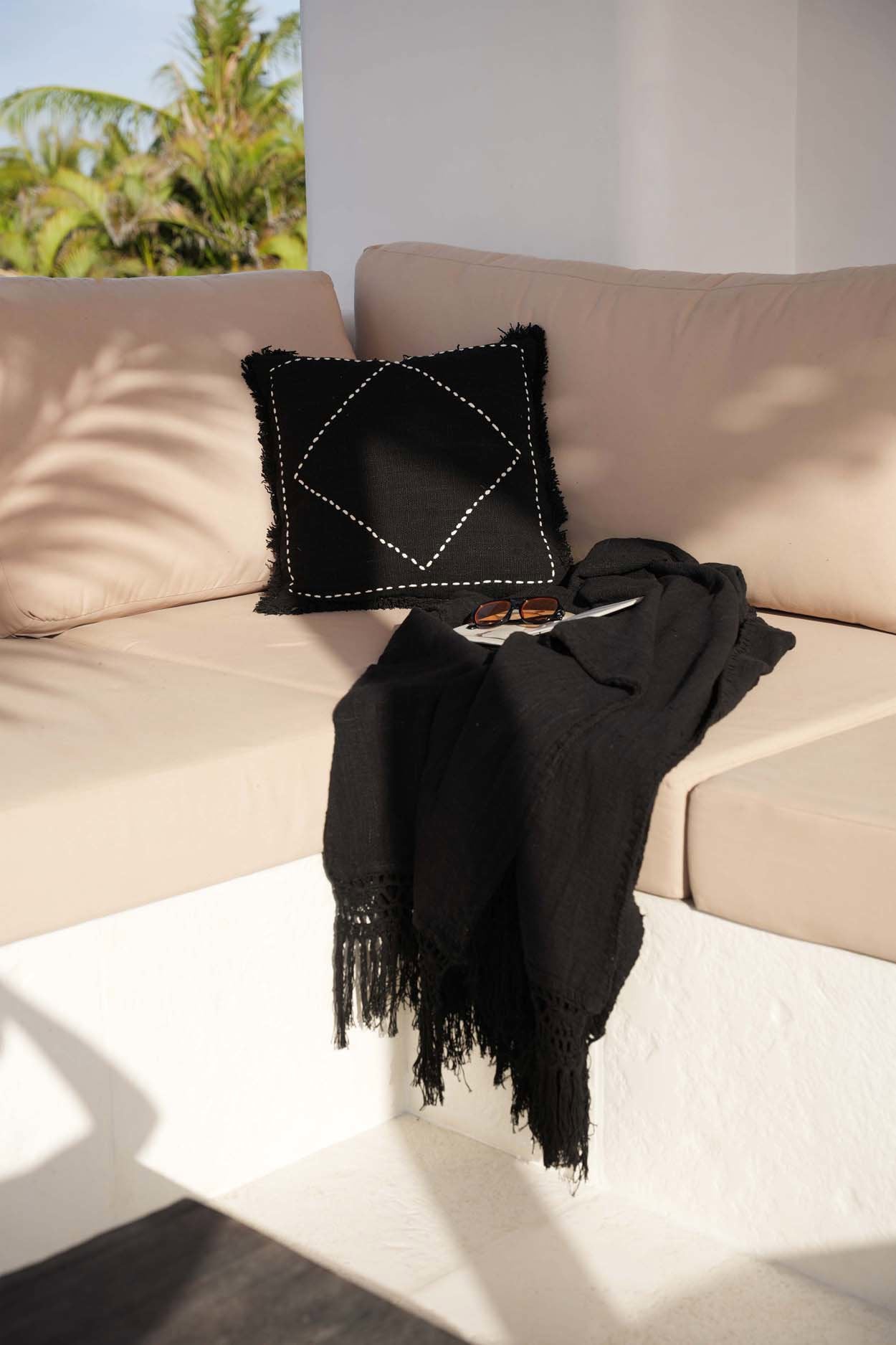 Kayu Living black cotton Gypsy bed throw with fringe detail draped on a beige outdoor sofa with a matching pillow.