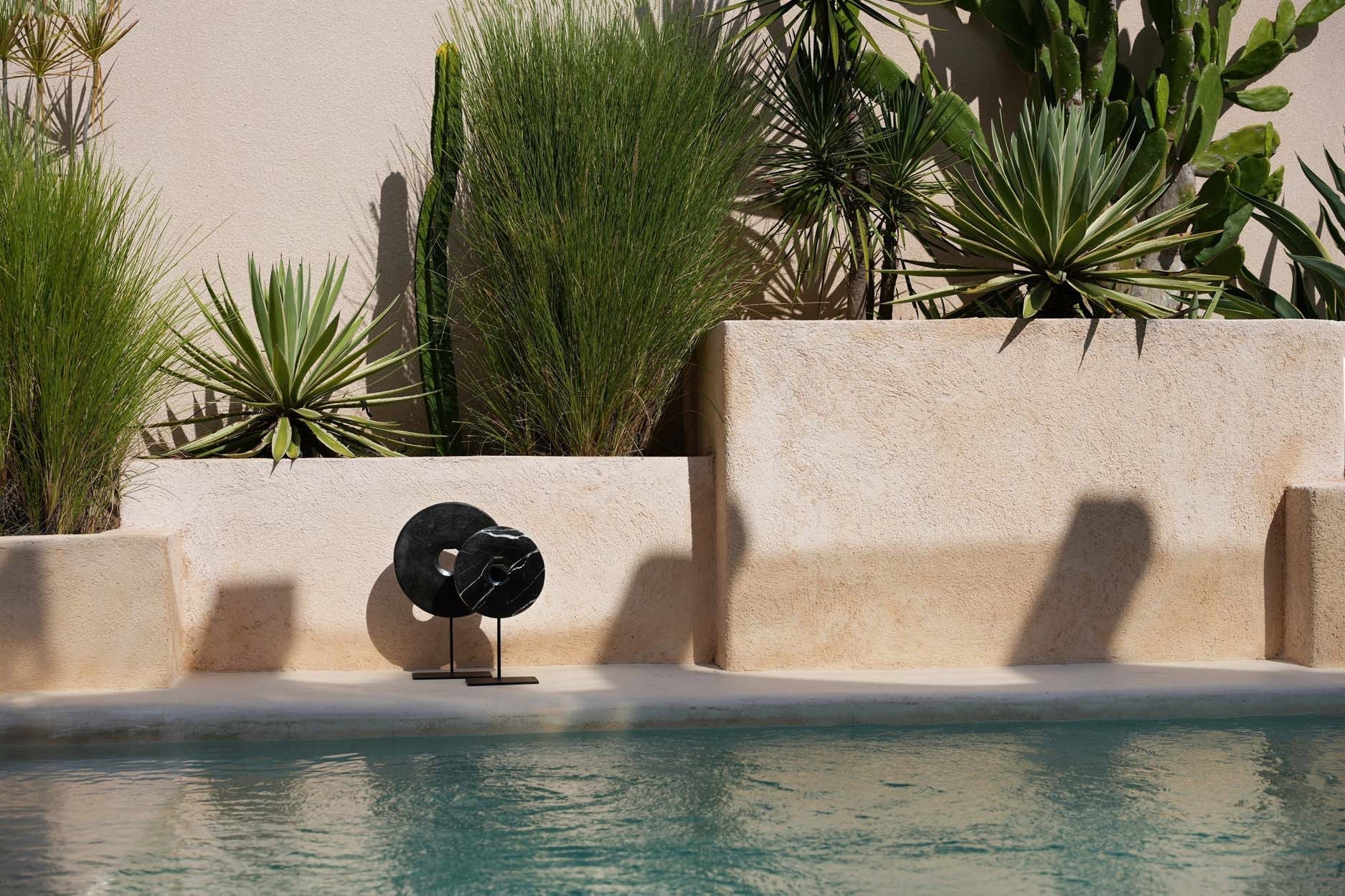 Kayu Living black marble disc sculpture on a metal stand displayed by a sunlit outdoor swimming pool.