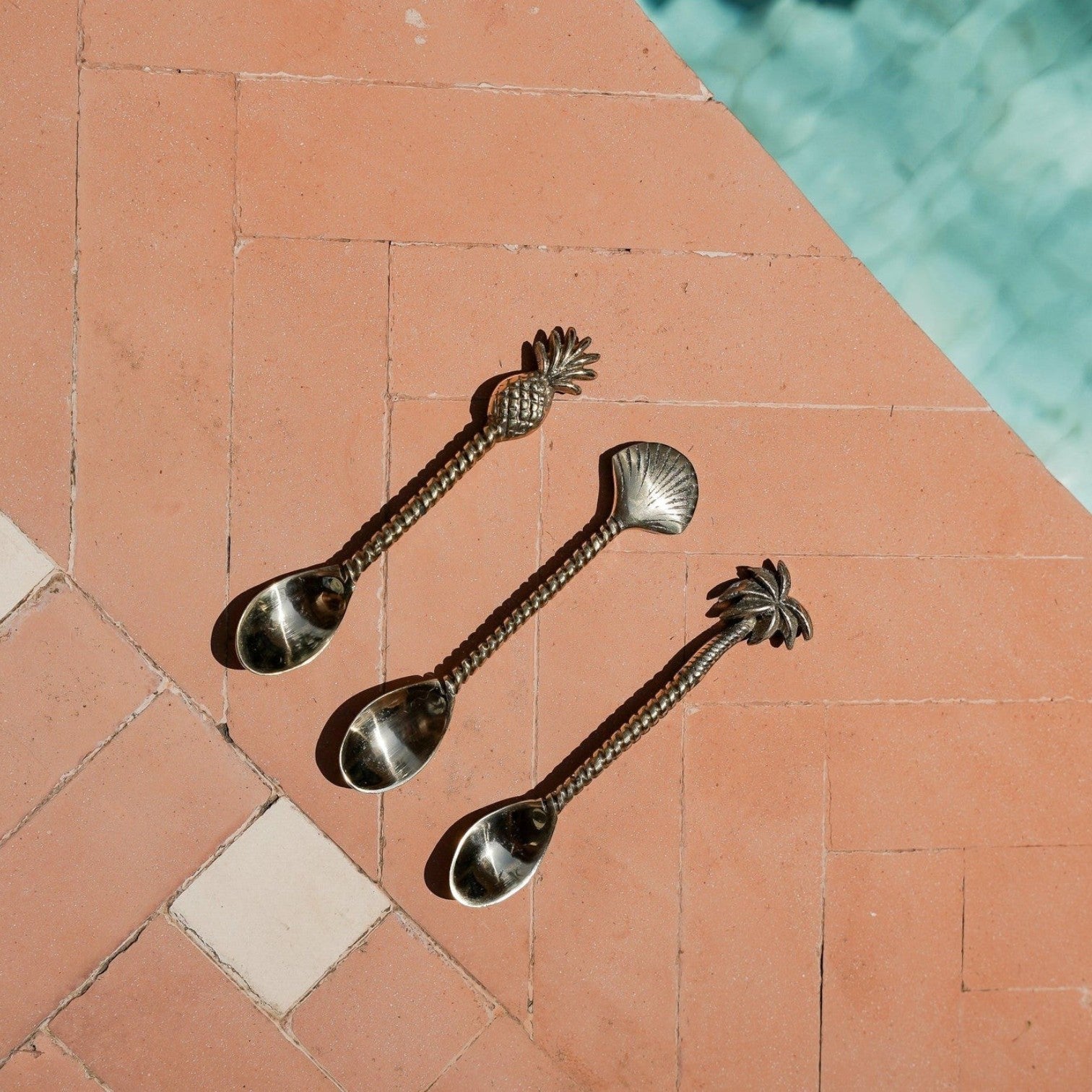 Kayu Living set of three handcrafted brass spoons featuring pineapple, shell, and palm tree motifs on a tiled patio.