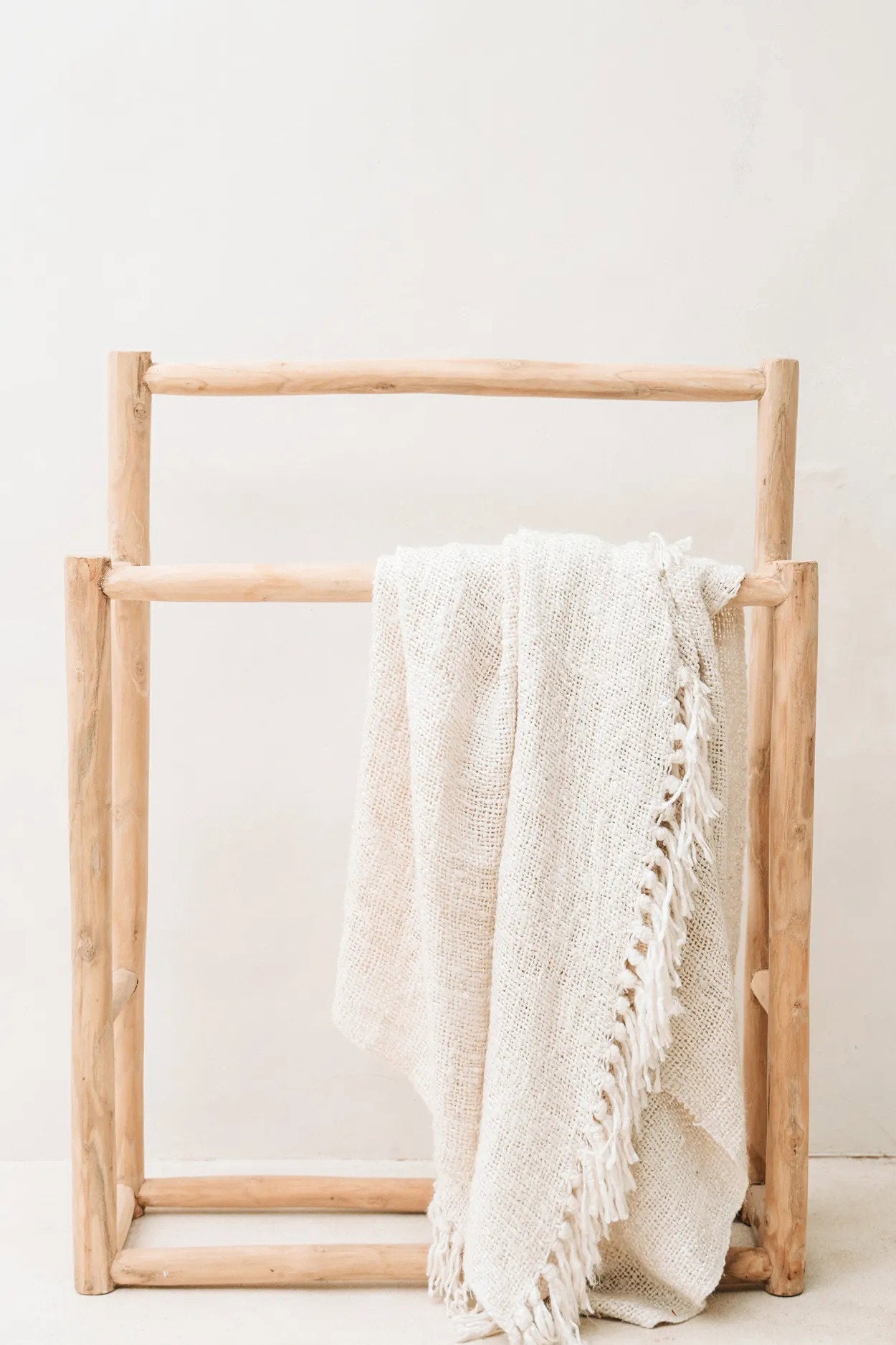 Kayu Living cream hand-woven cotton throw blanket with a waffled texture and fringe detail draped on a wooden rack.