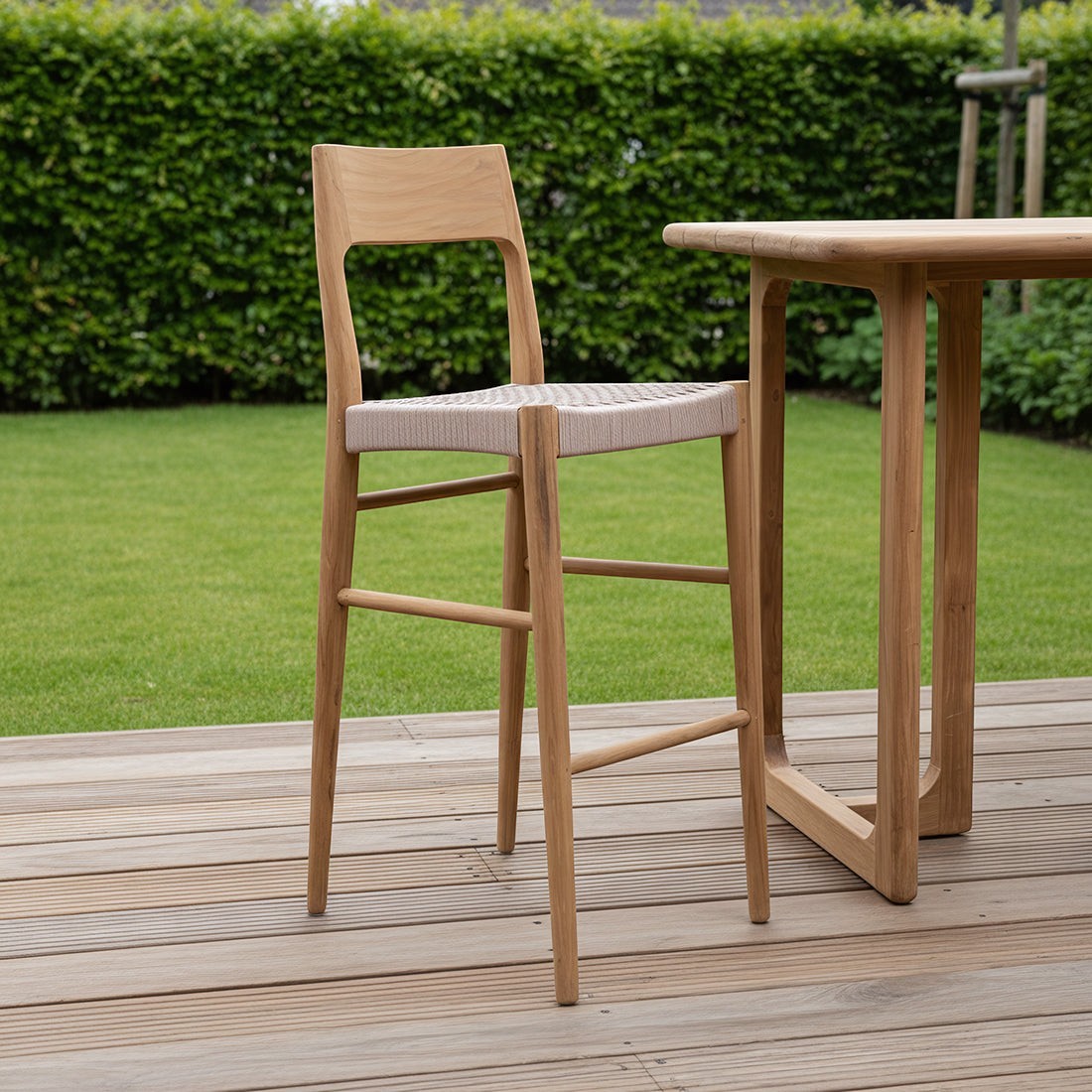 Kayu Living Freya bar stool in solid teak with a beige woven synthetic rope seat on a wooden outdoor terrace.