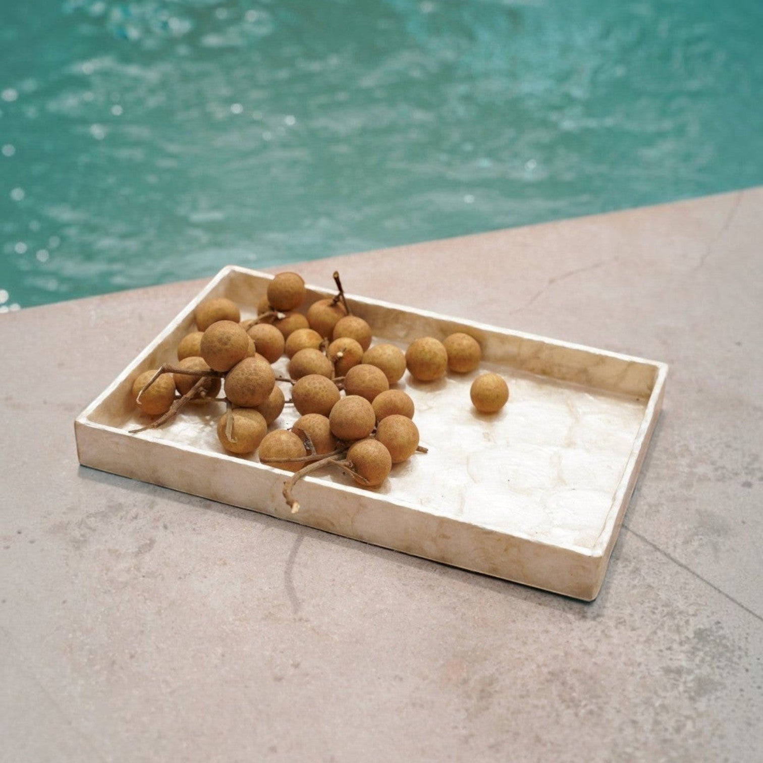 Kayu Living handmade gold Capiz shell rectangular tray holding longan fruit beside a swimming pool