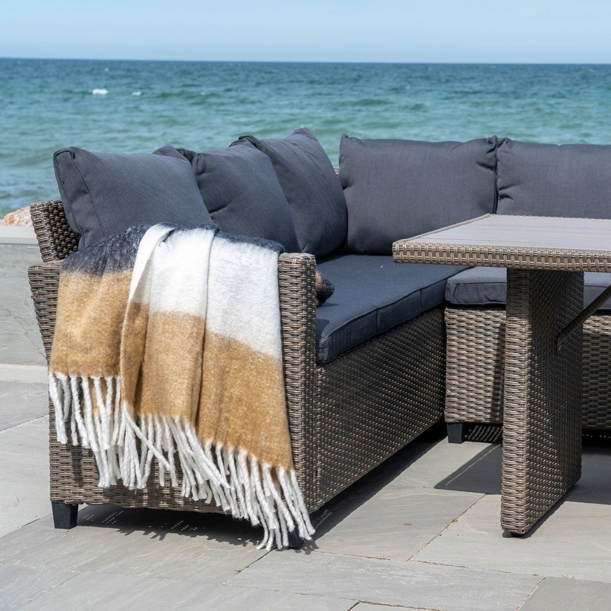 Kayu Living Sintra throw in black, white, and brown draped over a wicker outdoor sofa by the ocean.