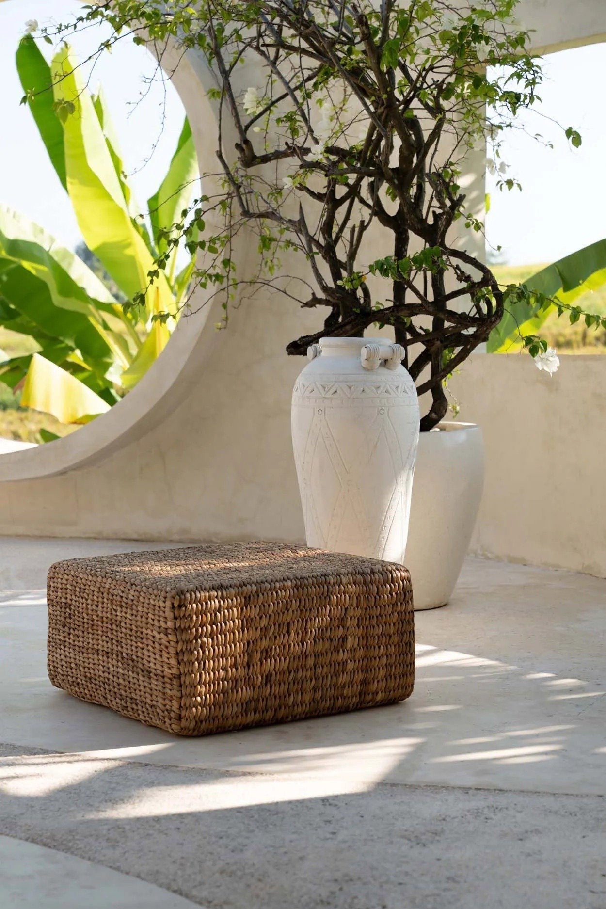 Kayu Living square water-hyacinth pouf in natural tan, set on a patio with a large white decorative vase.