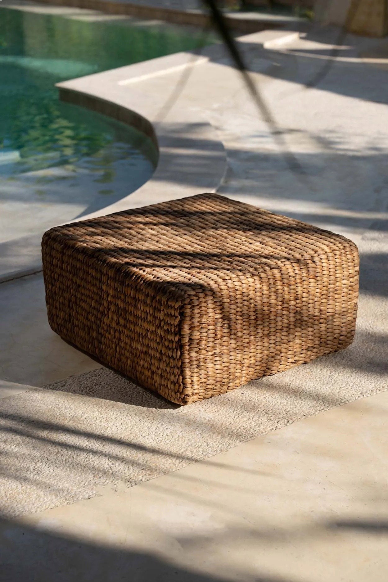 Kayu Living square water-hyacinth pouf in natural tan, featuring a woven texture on a poolside patio.