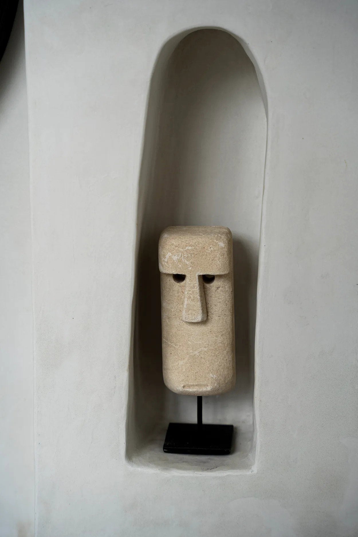 Kayu Living hand-carved sandstone Sumba statue on a black metal stand displayed in a white wall niche.