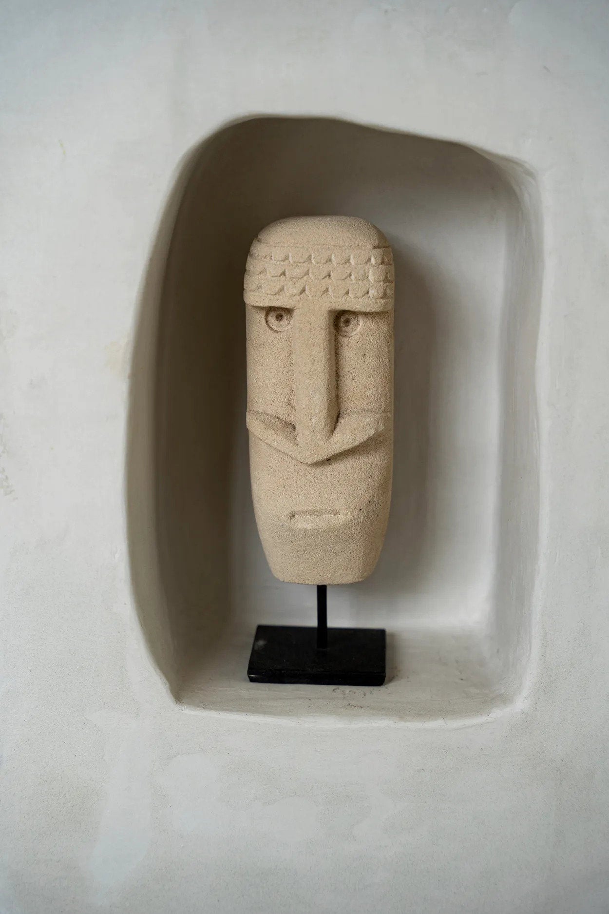 Kayu Living hand-carved beige sandstone Sumba statue on a black metal stand displayed in a white wall niche.