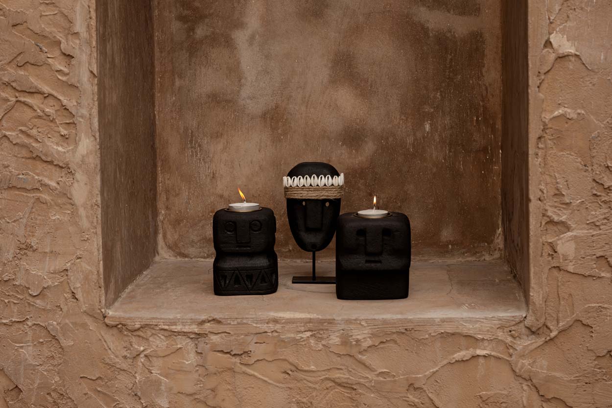 Kayu Living hand-carved black sandstone Sumba statues on stands with lit tea light candles in a rustic stone alcove.