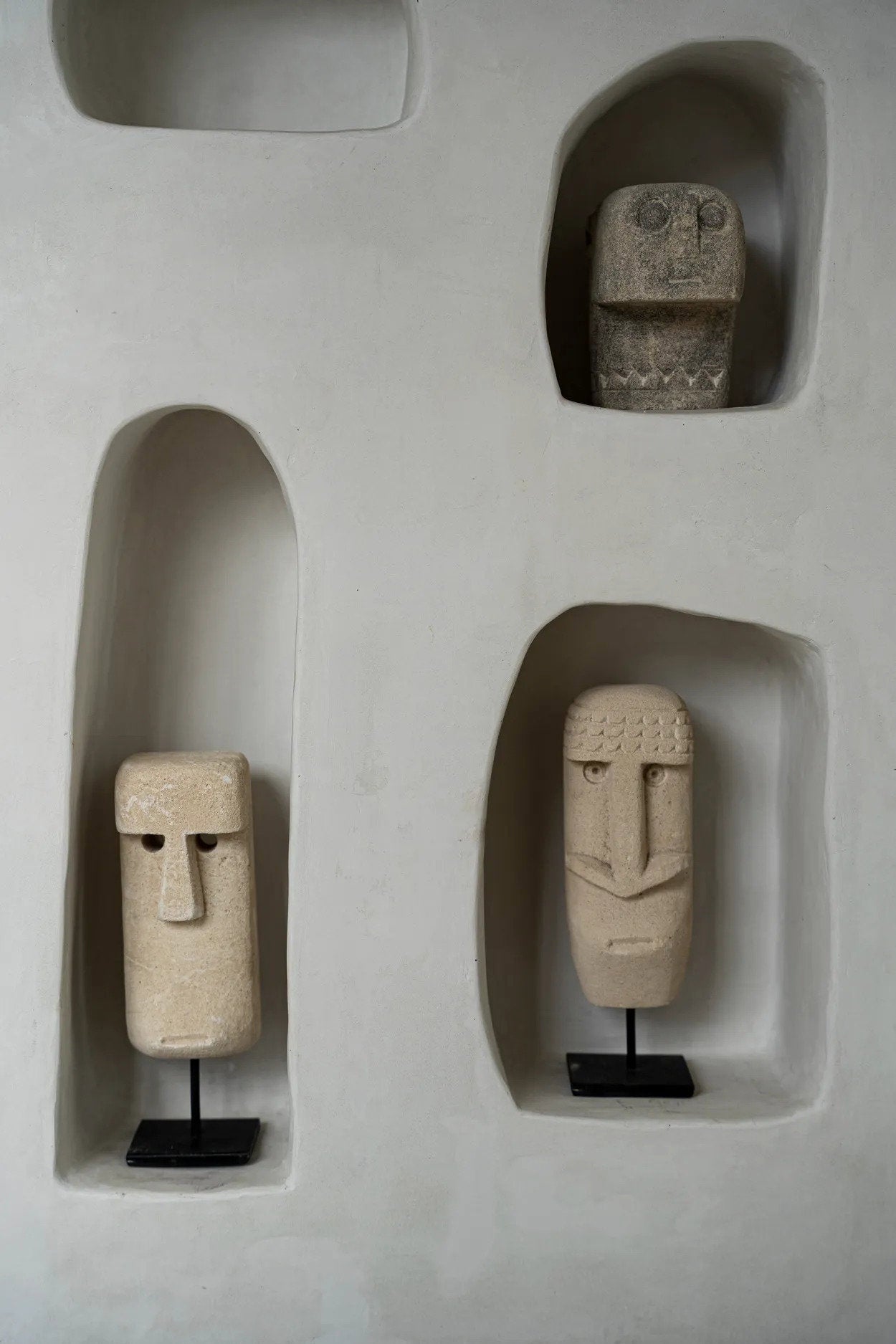 Kayu Living hand-carved sandstone Sumba stone statues on black metal stands displayed in white wall niches.