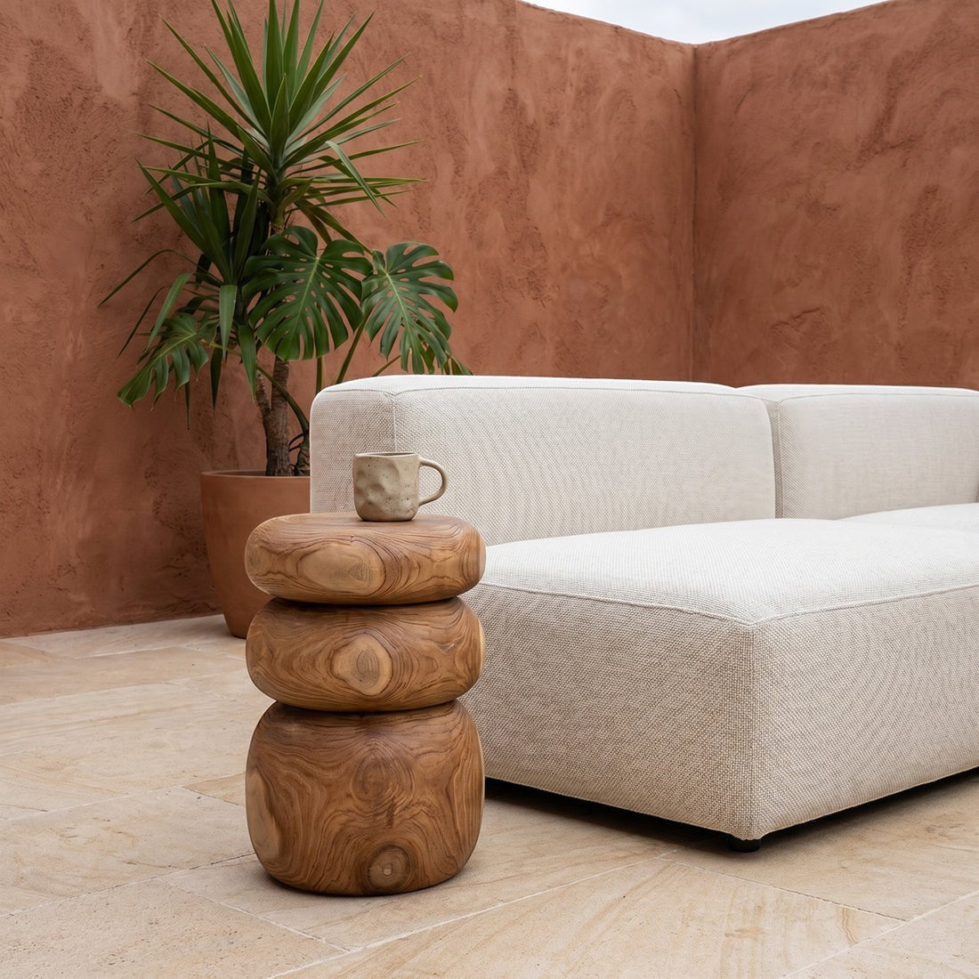 Kayu Living Tao solid teak side table with rounded stone-like shapes, placed on a terrace next to a beige sofa.