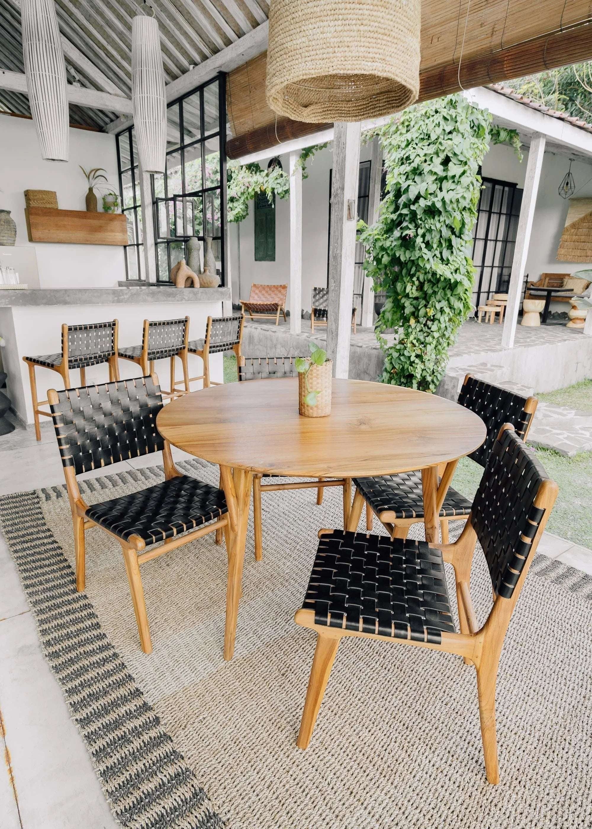 Kayu Living Pantai round 120 cm teak dining table with four black woven leather chairs. Natural wood grain.