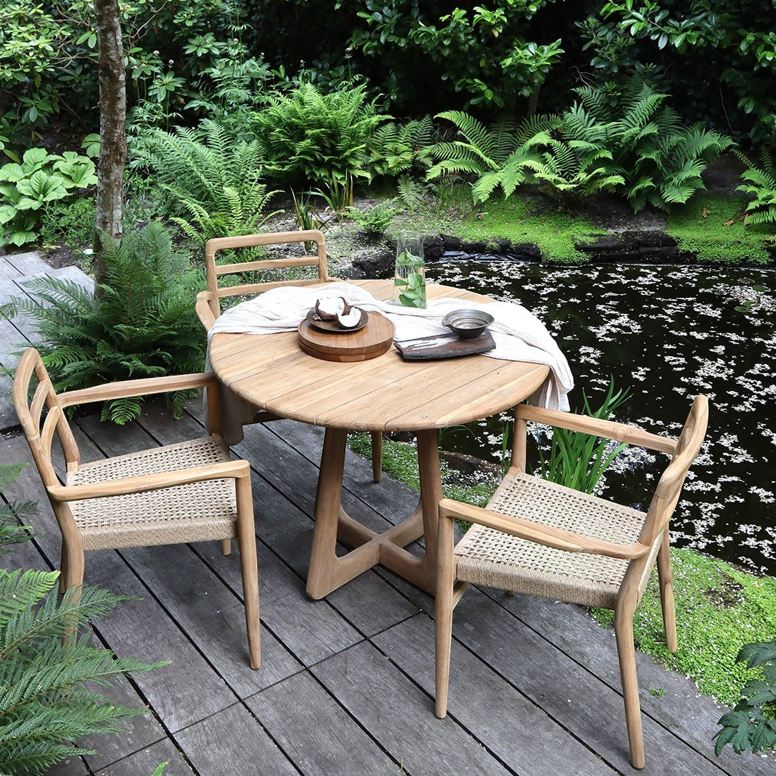 Kayu Living Verano round teak table with two woven chairs on a garden deck beside a pond.