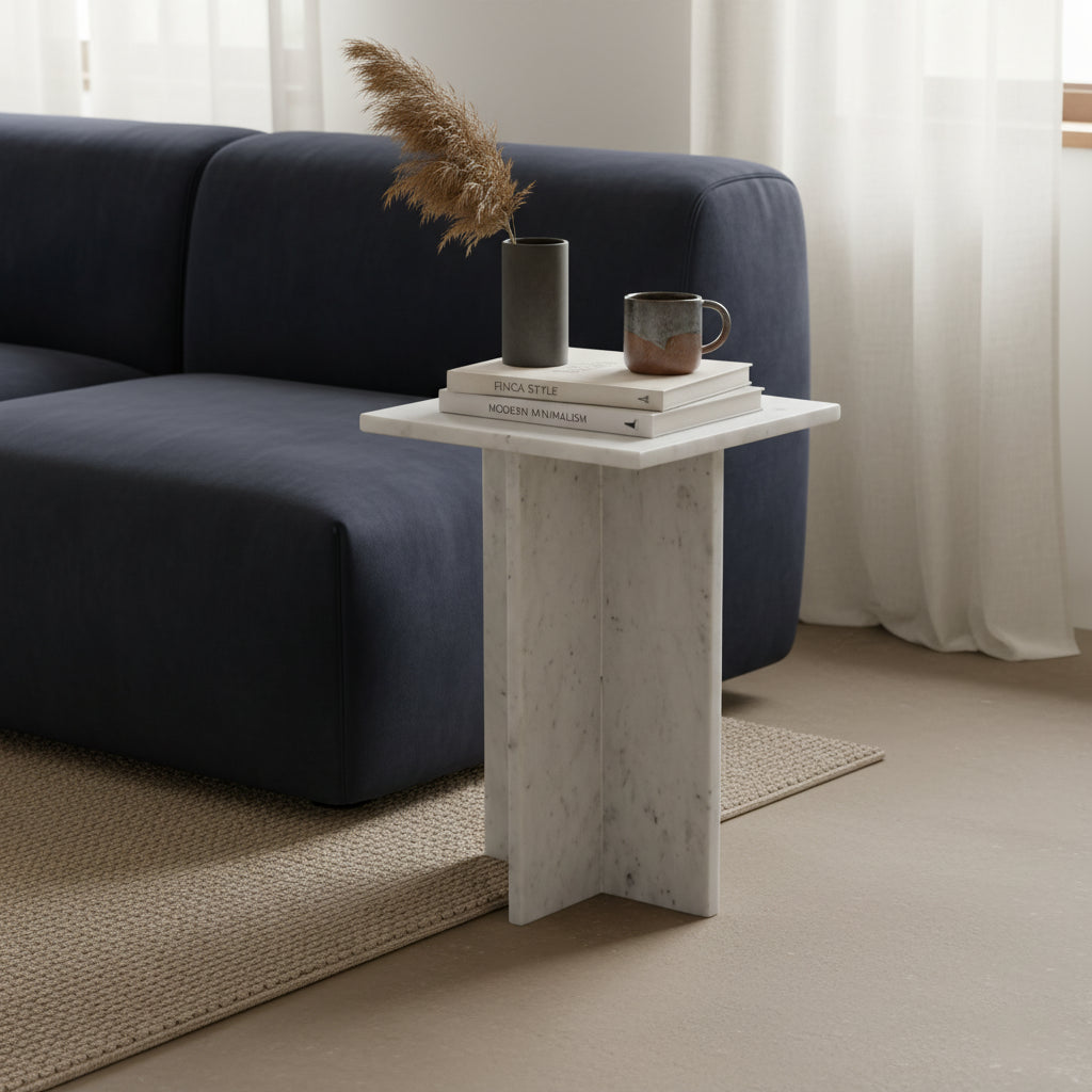 Kayu Living white marble side table with crossed slab base, styled with books and a vase next to a navy sofa.